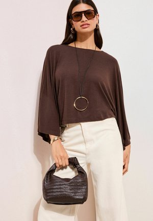 Brun langærmet top med vide ærmer, kombineret med off-white bukser med vide ben. Tilbehør inkluderer en sort flettet håndtaske og guldfarvet hoop halskæde.