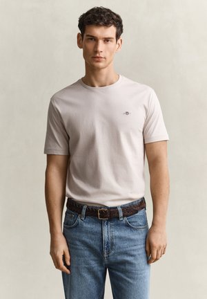 Jeune homme portant un t-shirt beige clair avec un petit logo sur la poitrine, un jean bleu et une ceinture en cuir tressé marron, debout devant un fond neutre.