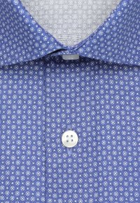Chemise bleu ciel avec un petit motif géométrique circulaire blanc, un bouton blanc fermé sous le col.
