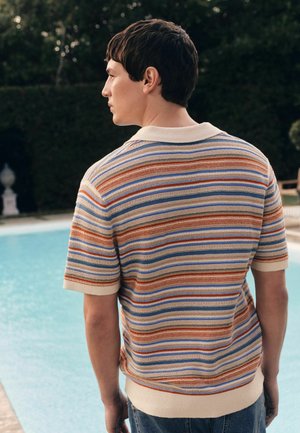 Tânăr bărbat într-o cămașă cu mâneci scurte cu dungi multicolore, stând lângă o piscină în aer liber, cu fața spre spate și capul întors spre stânga.