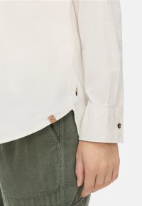 Witte langslievende shirt met een gestructureerde afwerking, met een zijsplit en een klein logolabel. Gecombineerd met groene ribstofbroek.