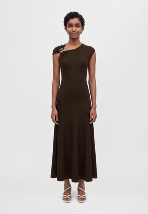 Une femme porte une robe marron foncé asymétrique à une épaule, longue jusqu’aux chevilles, avec une attache dorée sur l’épaule et des escarpins blancs à brides, sur un fond uni.