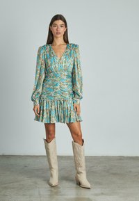 Mujer vistiendo un vestido mini fruncido de estampado azul con mangas largas y botas occidentales beige hasta la rodilla, de pie sobre un suelo gris.