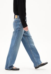 Jeans de pierna ancha en azul claro con un corte relajado, diseño de cinco bolsillos y material de denim, combinados con flats negros y una camiseta negra de manga larga.