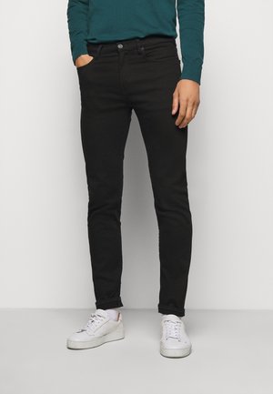 Homme portant un jean noir slim, des baskets blanches et une chemise à manches longues bleu sarcelle, une main dans la poche, debout devant un fond uni.