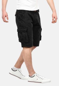 Black cargo shorts with multiple pockets, made of cotton fabric. Features include a relaxed fit and knee-length cut, paired with white sneakers.