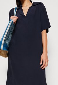 Robe courte bleu marine à manches courtes avec un décolleté en V et de subtils plis. Sac tissé bleu et beige avec bandoulière. Texture de tissu lisse.