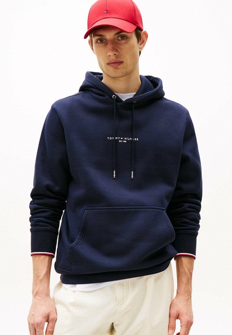 Felpa con cappuccio blu navy, dotata di tasca a marsupio e logo bianco. Accenti a strisce rosse e bianche sui polsini. Texture morbida e liscia.