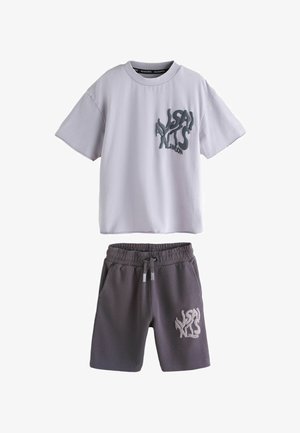 Fiú rövid, göndör hajjal, fehér, túlméretezett pólót visel, amelynek hátoldalán nagy fekete "ALLSAINTS LONDON" felirat található, szürke rövidnadrágot és fehér sportcipőt.