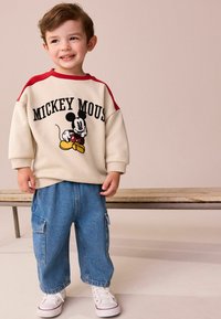 Crème sweatshirt met rode accenten met een Mickey Mouse grafiek, gecombineerd met blauwe denim broek en witte sneakers, staande op een houten bank.