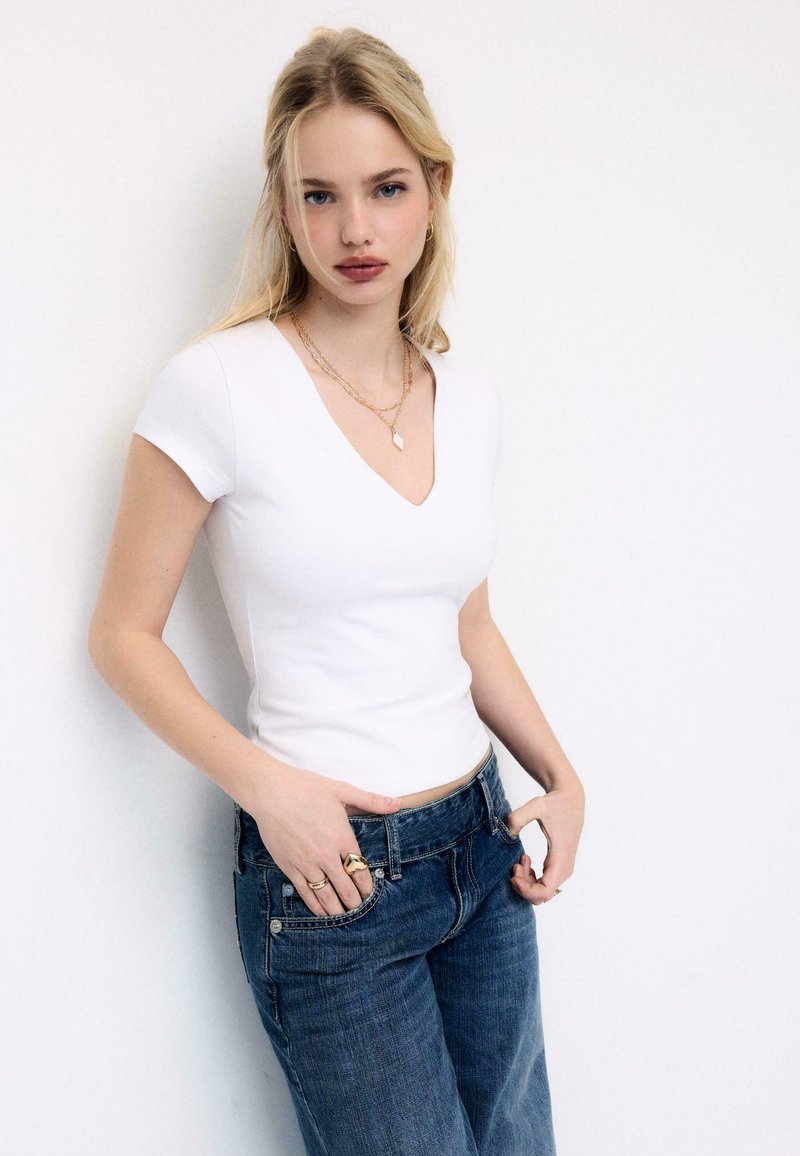Jeune femme aux cheveux blonds portant un t-shirt blanc à col en V, un jean bleu et des bijoux en or, debout contre un mur blanc.
