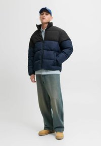 Czarna i granatowa puchowa kurtka z wysokim kołnierzem i zapięciem na zamek. Zestawiona z jasną koszulą w kratę, niebieskimi dżinsami i brązowymi chodakami.