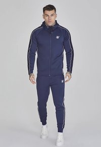 Granatowy dres z kurtką na zamek i spodniami, z białymi paskami po bokach i detalem logo. Wykonany z gładkiego materiału, dopasowany fason z kapturem.