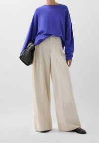 Paarse oversized sweatshirt gecombineerd met crèmekleurige wijde broek. De broek heeft zichtbare naden en het model houdt een zwarte leren tas vast.