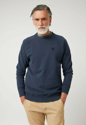 Sudadera azul marino con puños y dobladillo acanalados, cuello redondo y un pequeño logo circular en el pecho; combinada con pantalones de color beige claro.