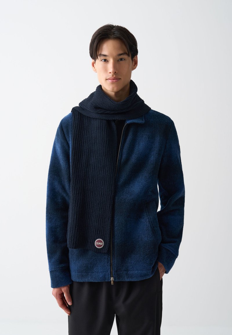 Jachetă din fleece bleumarin cu fermoar și eșarfă întunecată cu nervuri; are un logo rotund la tiv, țesătură texturată și două buzunare laterale.