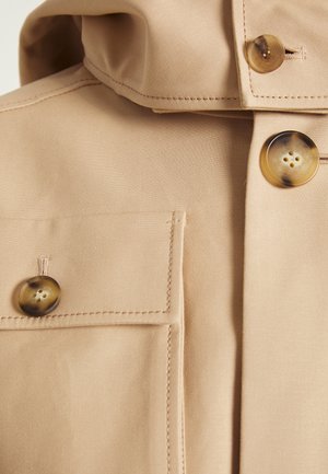 Col et poitrine de manteau en tissu beige avec de gros boutons en écaille et des détails de coutures visibles.