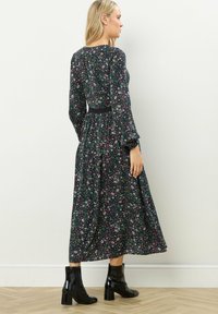 Vestido largo de manga larga con estampado floral en verde oscuro, que presenta una cintura ajustada y una falda de volantes. Botas negras hasta el tobillo complementan el conjunto.