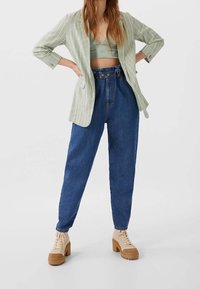 Helles grünes Blazer mit Nadelstreifen über einem grauen Cropped-Top, kombiniert mit hochtaillierten blauen Jeans und beigen Plateau-Stiefeln.
