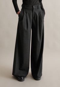 Wide-legbroek in donkergrijze stof met een gestructureerde afwerking. Voorzien van plooien aan de voorkant, riemlussen en zijzakken. Gecombineerd met zwarte schoenen.