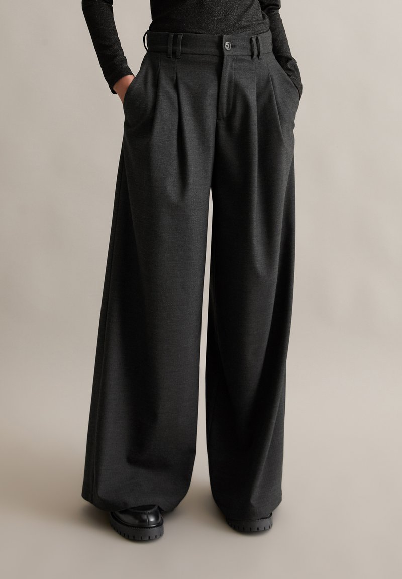Wide-legbroek in donkergrijze stof met een gestructureerde afwerking. Voorzien van plooien aan de voorkant, riemlussen en zijzakken. Gecombineerd met zwarte schoenen.