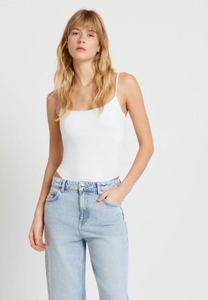 Giovane donna con capelli biondi lunghi che indossa una canottiera bianca e jeans azzurri a vita alta, mano in tasca, posa neutra.