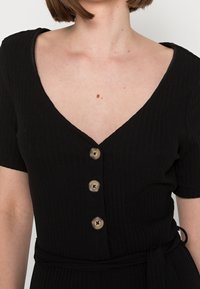 Czarna sukienka z prążkowanej tkaniny, z głębokim dekoltem w kształcie litery V, krótkimi rękawami i paskiem w talii. Posiada okrągłe brązowe guziki na przodzie. Gładka tekstura.