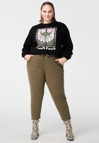 Femme debout, les mains dans les poches, portant un sweat-shirt noir avec un motif floral et le texte « Good Mood », un pantalon olive et des bottines en peau de serpent.
