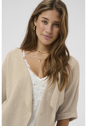 Giovane donna con capelli castano chiaro lunghi, che indossa orecchini a cerchio dorati, collane a strati, un cardigan beige e una maglia bianca in pizzo, che sorride dolcemente.