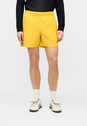 Osoba ubrana w żółte sportowe szorty, białe skarpetki i białe buty sportowe z czarnymi paskami stoi na tle jednolitego tła.