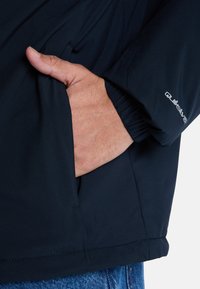 Mørkeblå jakke med en glat tekstur. Har en sidelomme og elastiske manchetter. Lille logo trykt på ærmet. Nærbillede af hånd i lommen.