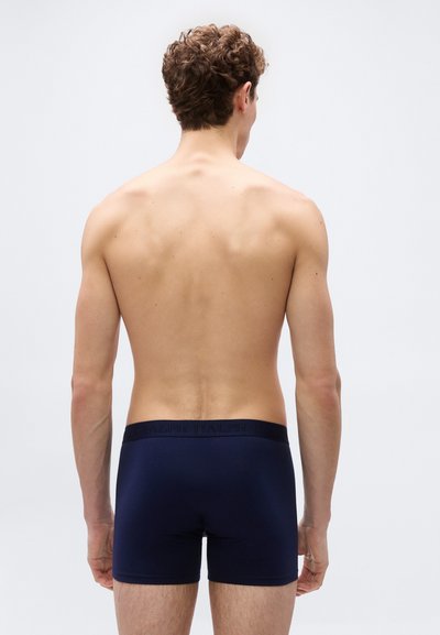 Άνδρας με σγουρά μαλλιά, χωρίς πουκάμισο και με navy μπλε μποξεράκια, στέκεται με την πλάτη προς το μέρος μας, σε απλό ανοιχτόχρωμο φόντο.