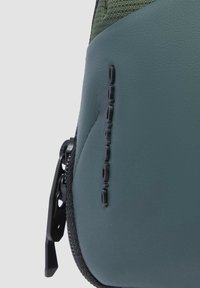 Bolsa verde oscura con la parte superior de tela texturizada y la sección inferior lisa. Cuenta con una cremallera negra y un logo de la marca en relieve en el lateral.