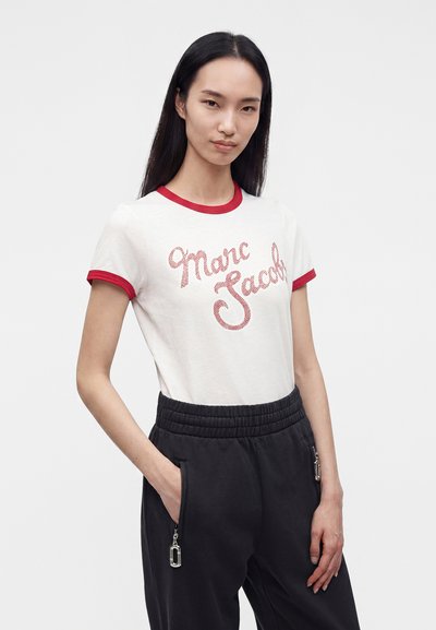 Γυναίκα με μακριά ίσια μαλλιά φοράει λευκό T-shirt Marc Jacobs με κόκκινο τελείωμα και μαύρο παντελόνι, στέκεται με το ένα χέρι στην τσέπη.
