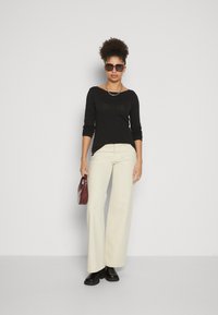 Haut noir à manches longues, pantalon large écru, bottines noires, lunettes de soleil marron, collier en chaîne argenté, sac à main bordeaux.