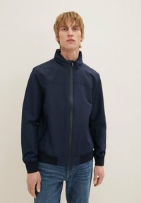Chaqueta ligera de color azul marino con un cuello alto, cremallera frontal y puños elásticos. Presenta bolsillos laterales y una textura suave y mate.