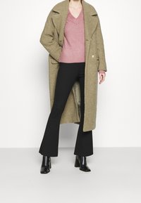 Olivgrön överdimensionerad kappa, rosa V-ringad tröja, svarta vida byxor och svarta klackstövlar. Mjuka texturer och knappdetaljer på kappan.