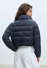 Námornícka puffer bunda s vysokým golierom, prešívanou textúrou, skráteným dizajnom a elastickým trojdielnym sťahovaním na spodnej časti. Biele nohavice v pozadí.