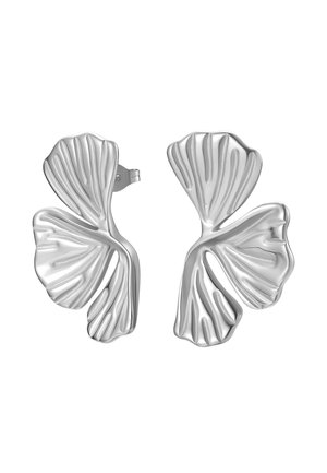 Pendiente de botón plateado con forma de doble hoja ondulada en la oreja izquierda, con la mano tocando suavemente el lóbulo de la oreja, cabello oscuro recogido por una diadema beige.