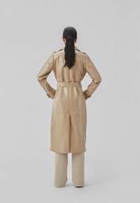 Beige frakke med en blank finish, med en bælte i taljen, skulderlapper og lange ærmer. Sammen med beige lige bukser.