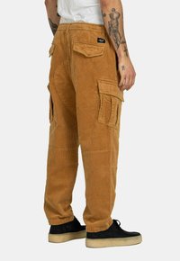 Corduroy cargo pants in tan with multiple pockets, a relaxed fit, and a textured surface. Worn with black shoes and a white shirt.