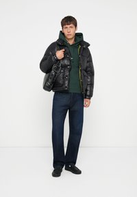 Chaqueta acolchada negra con un acabado brillante, sudadera con capucha verde, jeans azul oscuro y zapatos negros. Los accesorios incluyen un bolso negro de hombro.