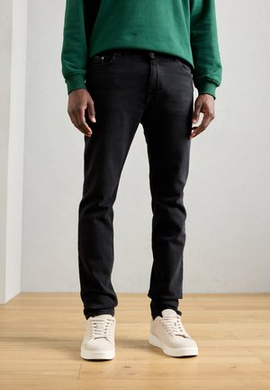 Zwarte spijkerbroek van denim, slim fit. Gecombineerd met beige sneakers, staand op een houten vloer tegen een neutrale muur. Groene sweatshirt erboven.