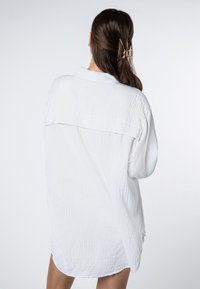 White, textured oversized shirt with a button-down front and back pleat detail, featuring a relaxed fit and curved hem.