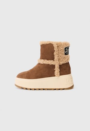 Stivale alla caviglia in suede marrone con bordi in shearling beige, caratterizzato da una suola piatta bianca spessa e testurizzata e un'etichetta con logo nera sul lato.