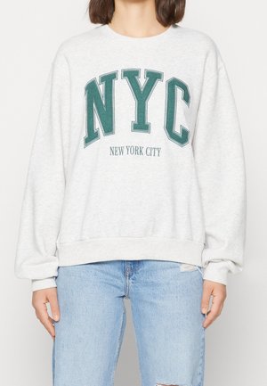 Person iført en lysegrå sweatshirt med store grønne "NYC" bogstaver og teksten "New York City", kombineret med lyseblå, revnede jeans.