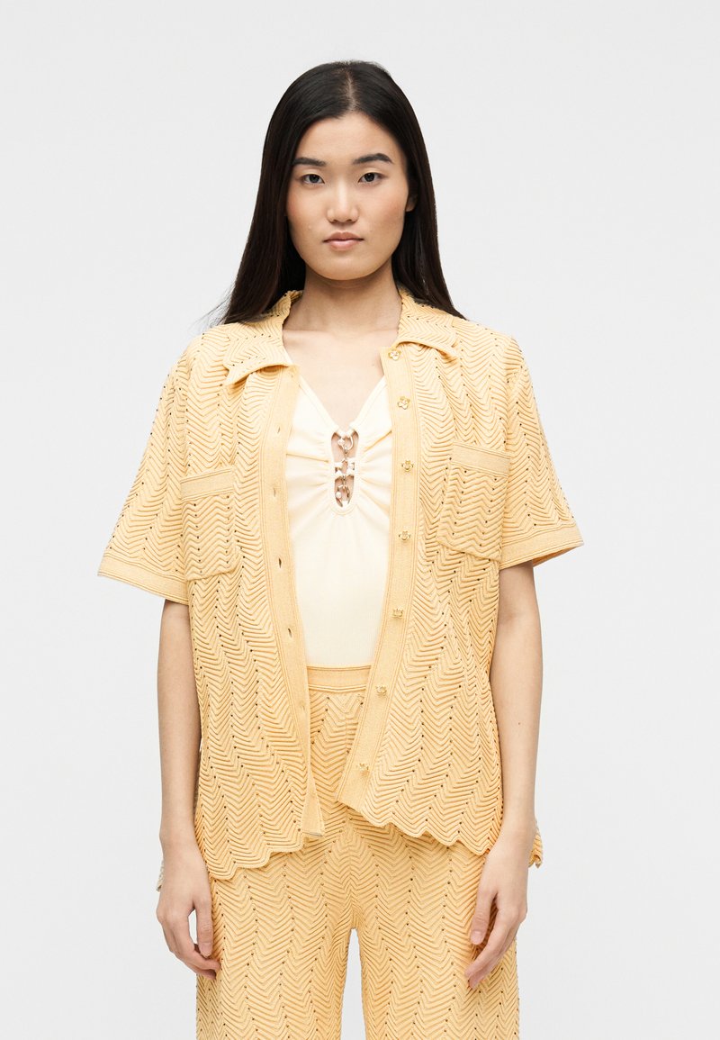 Γυναίκα με μακριά σκουρόχρωμα μαλλιά φορά ένα ζευγάρι ροδακινί πλεκτό κοντομάνικο μπλουζάκι και παντελόνι με υφή, πάνω από εκρού τοπ με μεταλλικούς κρίκους.