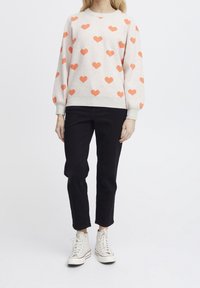 Sweat-shirt gris clair avec motif cœur orange, poignets et ourlet côtelés, associé à un pantalon noir et des baskets montantes blanches.