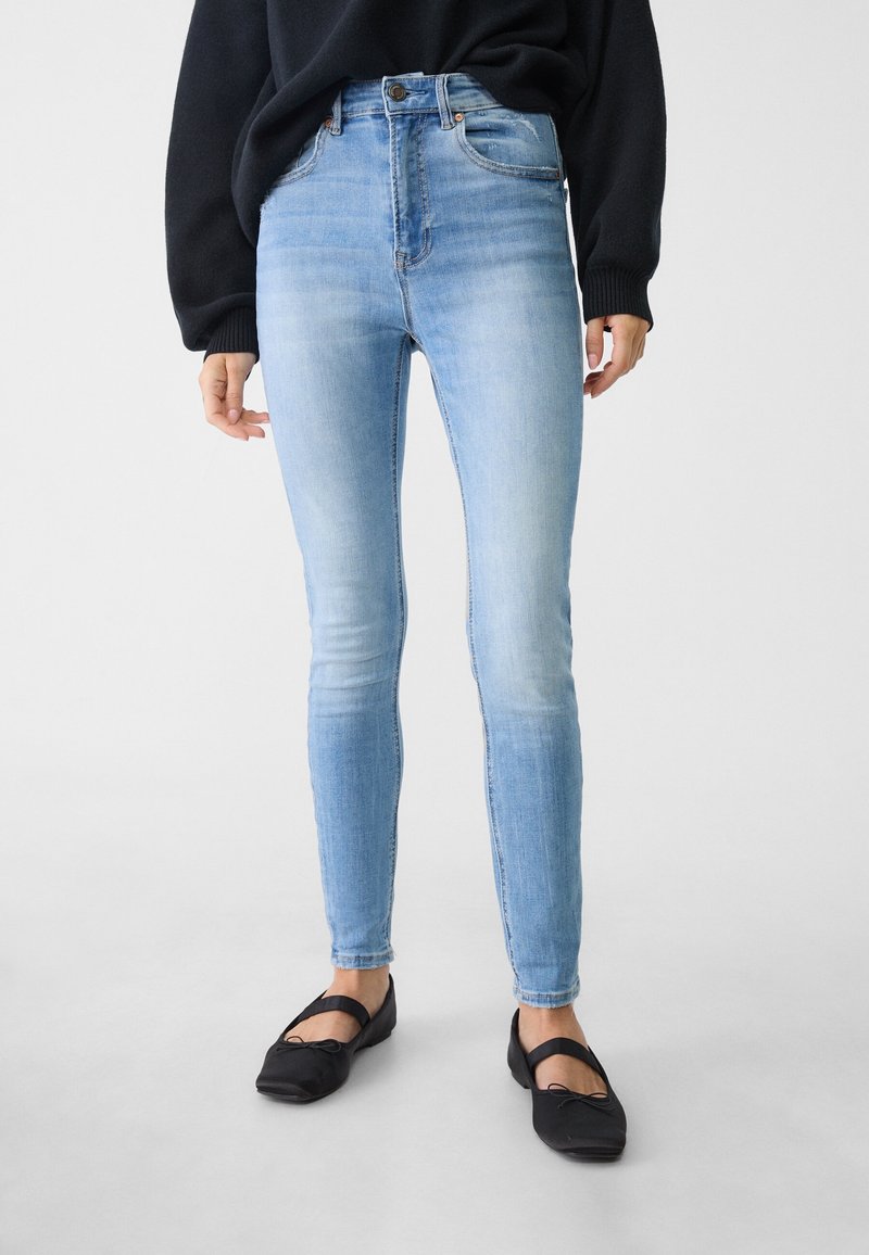 Jeans skinny de mezclilla azul claro con cintura alta, que presentan un ligero desgastado y un cierre de botón en la parte delantera. Unos zapatos planos negros complementan el atuendo.