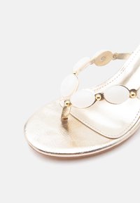 Χρυσό σανδάλι με λευκά οβάλ διακοσμητικά και μικρά χρυσά καρφιά, με θηλιά για το δάχτυλο και ανοιχτή μύτη σε λευκό φόντο.